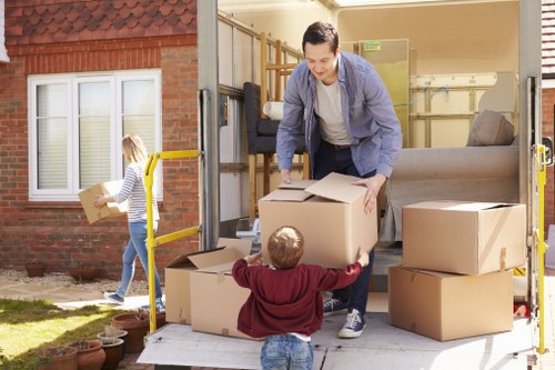 A man wearing a grey jacket and light trousers is seen loading large cardboard boxes into the back of a moving van parked outside a residential house in Harringay. The van's rear doors are open, revealing a partially filled interior with additional boxes and furniture, including a grey sofa. A young girl with blonde hair, dressed in a striped top and jeans, is walking away from the van while carrying a cardboard box. The scene takes place on a sunny day in a suburban street with a brick house featuring white-framed windows and a small garden with potted plants visible in the background. The man is carefully placing a box onto the van's loading area, with other packing boxes stacked nearby, suggesting an organized move typical of professional house removals services in the Harringay area. The overall setting emphasizes an active residential move involving household items like boxes and furniture, with a focus on careful handling and outdoor loading activities related to house relocation.