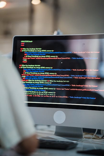 Close-up image of a laptop on a desk displaying a code editor with colorful syntax highlighting, including HTML and CSS code, alongside a blurred background with a mobile phone. The laptop keyboard is