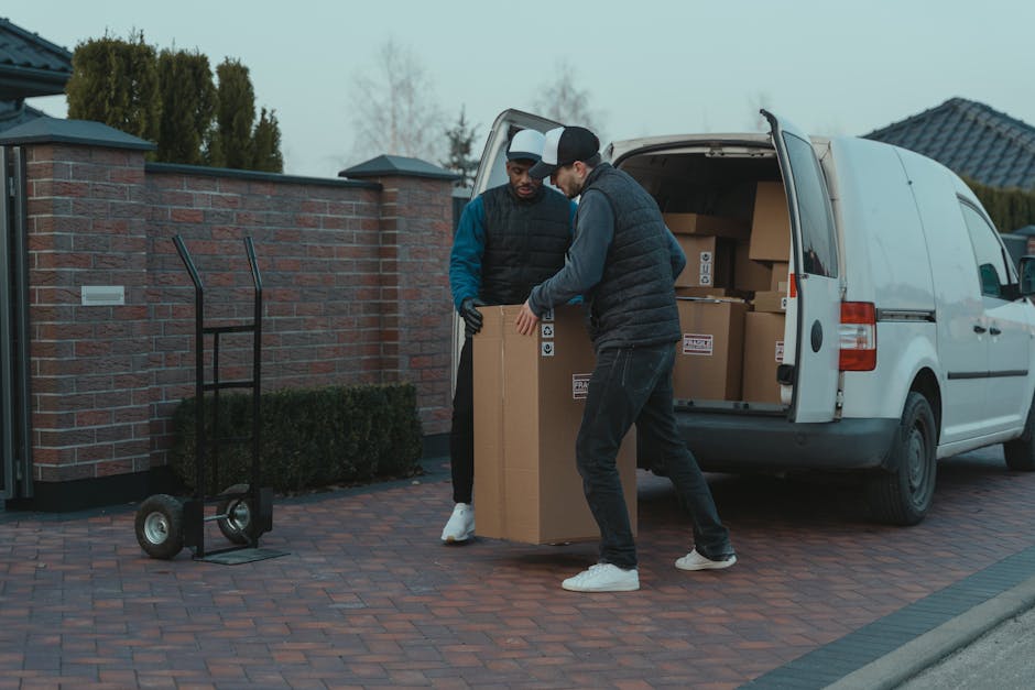 Two movers dressed in dark uniforms are seen loading a large, dark green upholstered piece of furniture into the open rear of a white van parked on a street, with both individuals lifting the item car